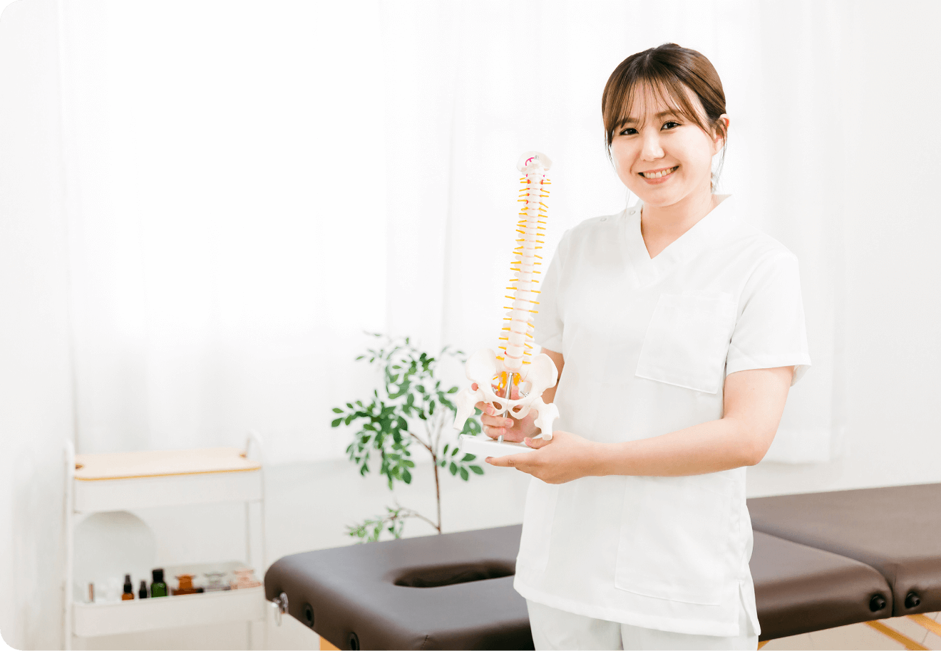 施術ベッドの前で脊椎模型を持って説明する笑顔の女性スタッフ