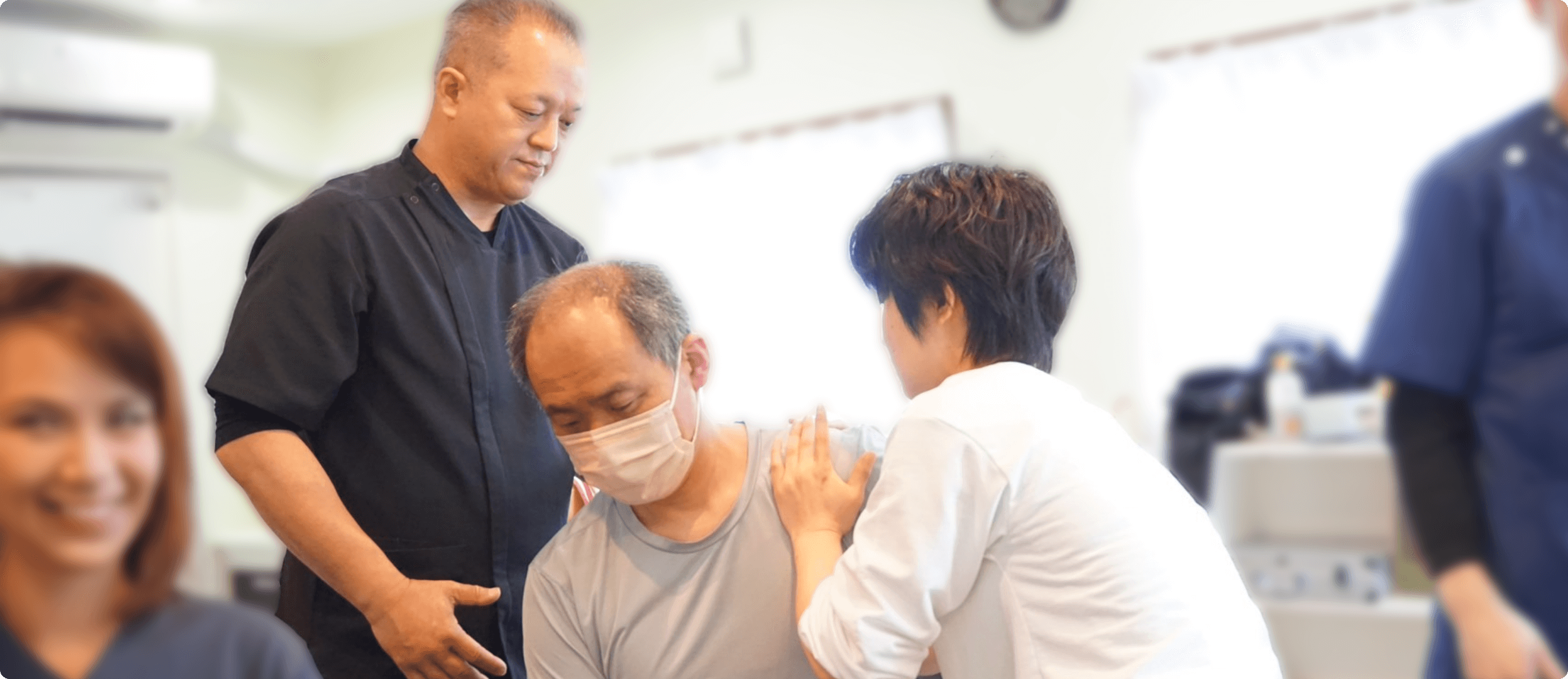 数人の施術者が集まり、高齢男性に対してリハビリの指導やサポートをしている場面