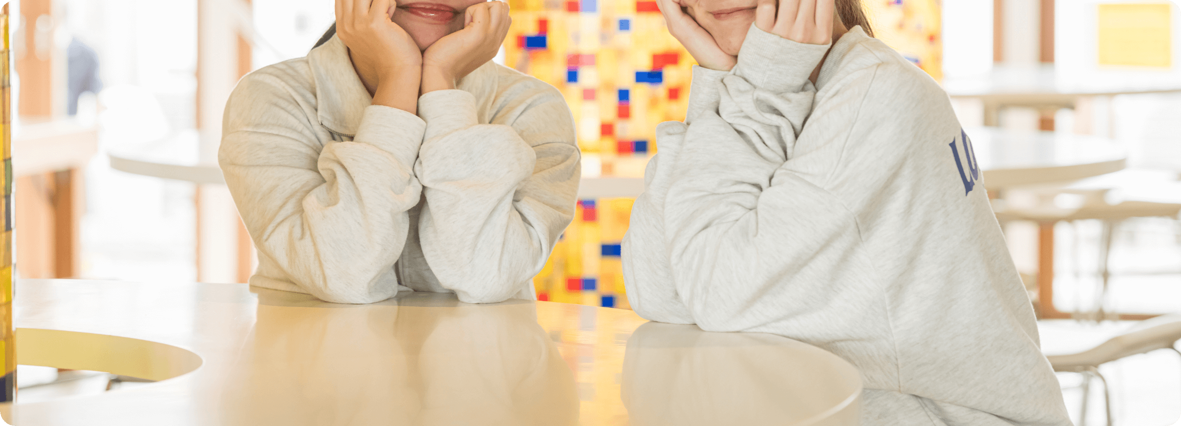 明るい室内で、二人の若い女性が並んで両手で頬杖をつき、笑顔を見せている。