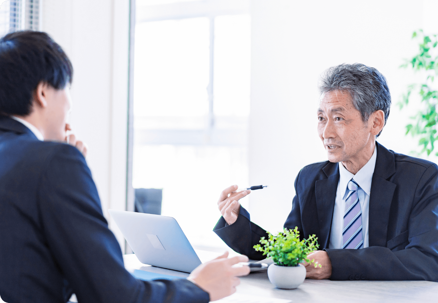スーツ姿の男性二人がオフィスの机越しに向かい合い、会話をしている場面。
